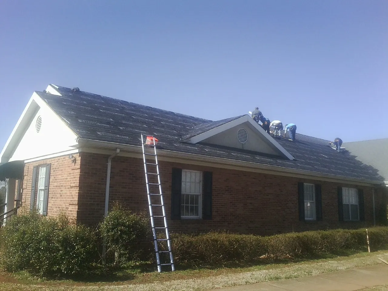 Crew tearing shingles off a brick ranch roof with a ladder set — Metal Roof Repair work by our crew in Knob Noster